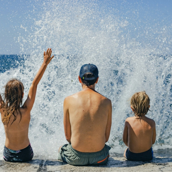 Das Bild zeigt drei Kinder, die mit dem Rücken zur Kamera auf einer niedrigen Steinmauer oder Uferkante sitzen. Sie blicken auf das Meer.