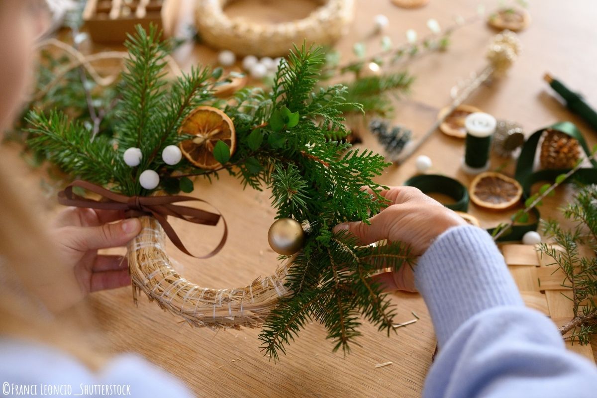 Frau bastelt einen Weihnachtskranz mit Tannenzweigen, getrockneter Orange, Schleife und Weihnachtskugel